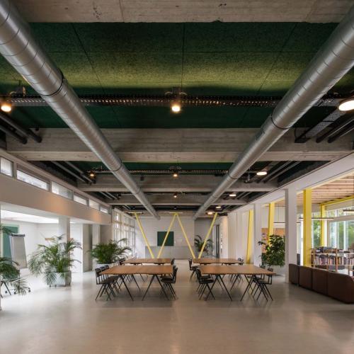 Modern binnenruimte met lange tafels en veel planten, gekenmerkt door een donkergroen plafond met zichtbare, zilveren leidingen.
