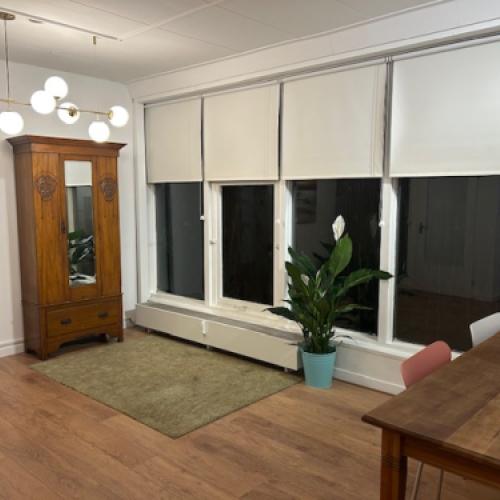 Lichte woonkamer met ramen voorzien van witte rolluiken en een houten kledingkast.