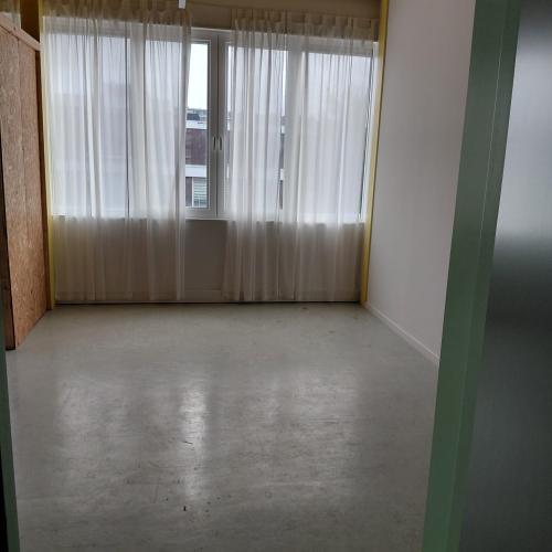 Interieur met grote raampartij voorzien van licht doorlatende gordijnen, een houten plafond en een lege, kale vloer.