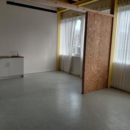 Een lichte, kale ruimte met een kastenwand, grote ramen met witte gordijnen en een zichtbaar houten afscheidingspaneel.