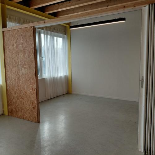 Een leeg, minimalistisch interieur met grote ramen, een houten scheidingswand, en een radiator aan de rechtermuur.