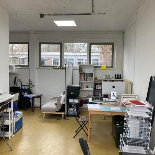 Binnenbeeld van een lichte, grote studio of werkplaats met ramen, diverse werkbanken en opgestapeld materiaal.