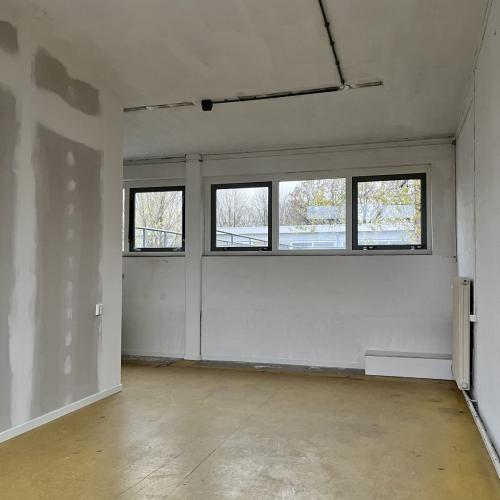 Groot, leeg interieur met lichtgekleurde muren, een glieven vloer en drie ramen in de achterwand.