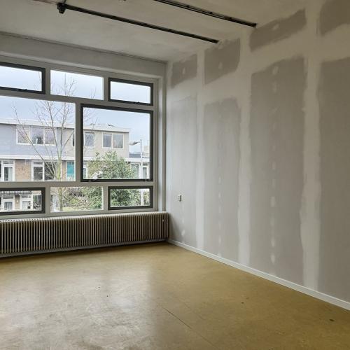Leeg en fel ingevallen interieur met grote raampartij en radiator; de muren hebben stucwerksporen.
