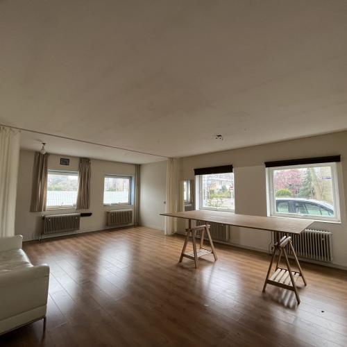 Lichte woonkamer met houten vloer, bank, ramen en een lang, robuust eettafelblad.