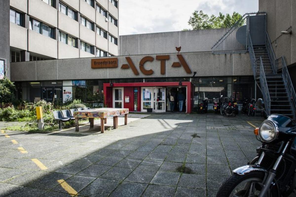 Vooraanzicht van een winkel (Acta) met een parkeerplaats ervoor, naast een betonnen gebouw.