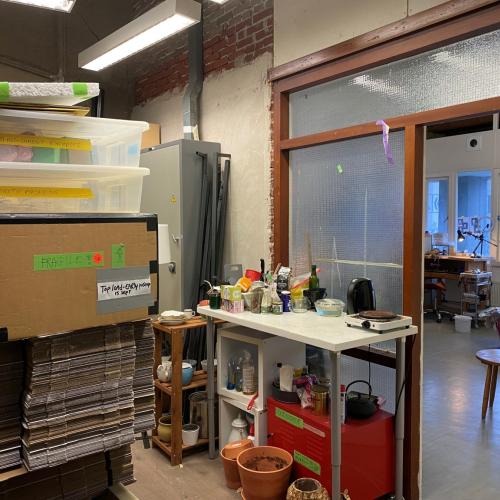 Interieur van een winkel met gestapelde goederen links, een toonbank met producten, en een glazen doorgang naar het achtergedeelte.