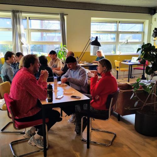 Interieur met een groep mensen die samenkomen in een lichte, moderne ruimte; er staan tafels en stoelen.