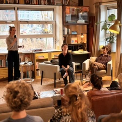 Interieur met een grote raampartij, boekenkasten en mensen die zittend in een salon praten.