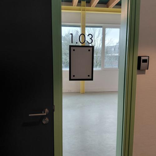 De deuropening leidt naar kamer 1.03, gemarkeerd met een nummerbord. Het zicht is licht door grote ramen.