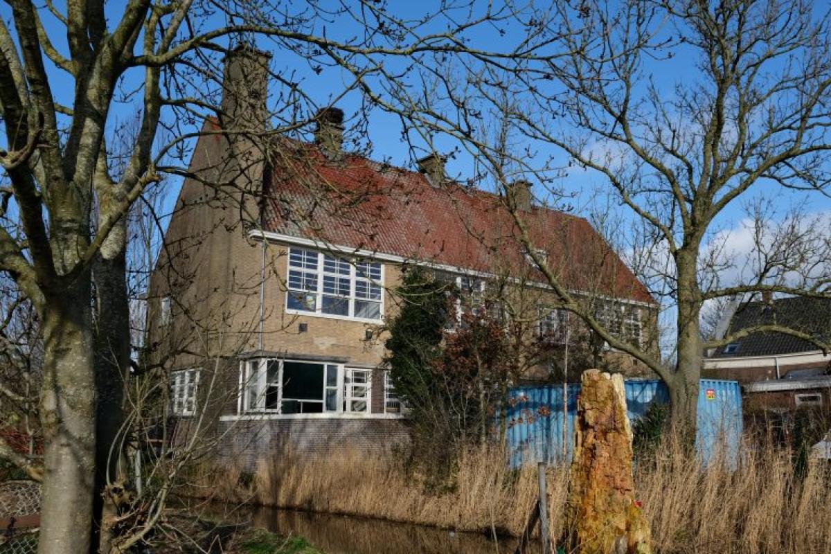 Oud, bakstenen huis met oranjerood dak, omgeven door bomen en bij een gracht.