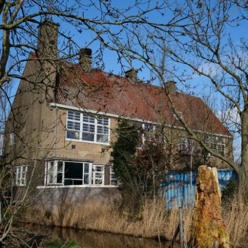 Oud, bakstenen huis met oranjerood dak, omgeven door bomen en bij een gracht.