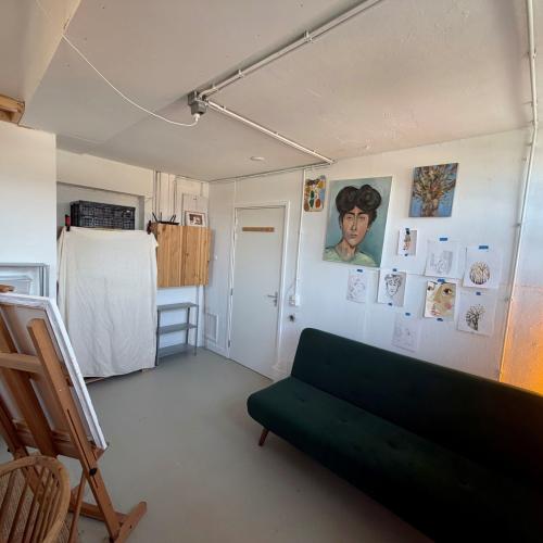 Studiointerieur met een schildersezel, een donkergroene bank en diverse ingelijste kunstwerken op de lichte muur.