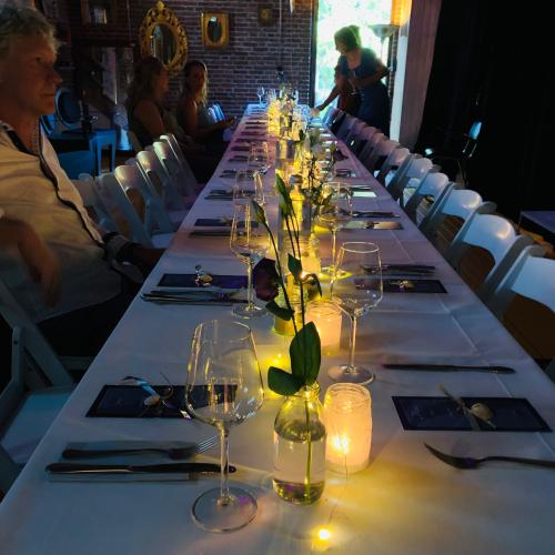 Lange tafel gedekt voor diner, versierd met kaarsen, bloemen en groene decoraties.
