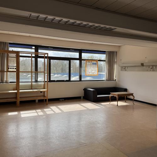 Ruime binnenruimte met grote ramen, een houten plankenopstelling links en een modern donker zitgedeelte met een tafel rechts.