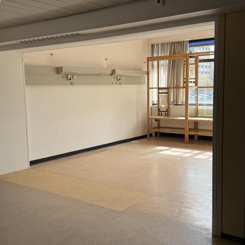 Lichte, lege kamer met een grote witte muur en een raam met houten scheidingswand rechts.