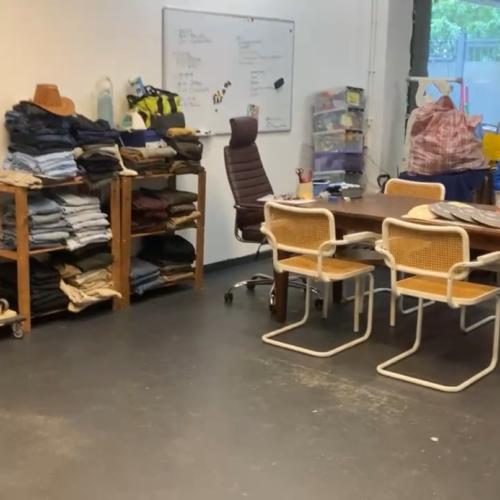 Kledingrekken met tweedehands kleding, gestapelde spullen en een houten tafel met stoelen in een grote verkoopruimte.
