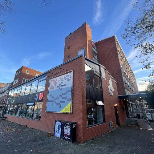 Straatbeeld van bakstenen gebouwen; een modern, laag gebouw met grote ramen domineert de voorgrond en staat naast een oude gevel.