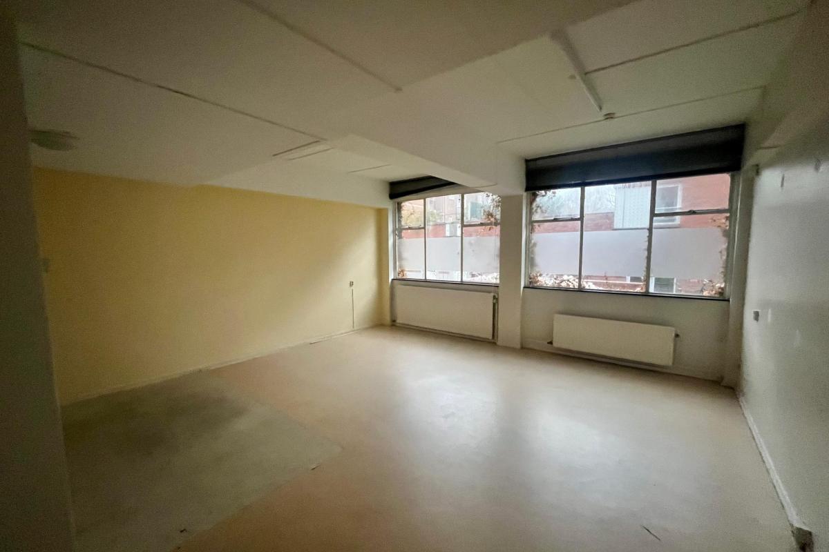 Lege, lichte kamer met onafgewerkte vloer, grote raampartij en lege muren.