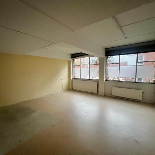 Lege, lichte kamer met onafgewerkte vloer, grote raampartij en lege muren.
