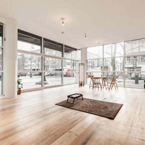 Ruime, moderne leefruimte met houten vloer en grote ramen met straatuitzicht. Centraal staat een donker vloerkleed en meubels.
