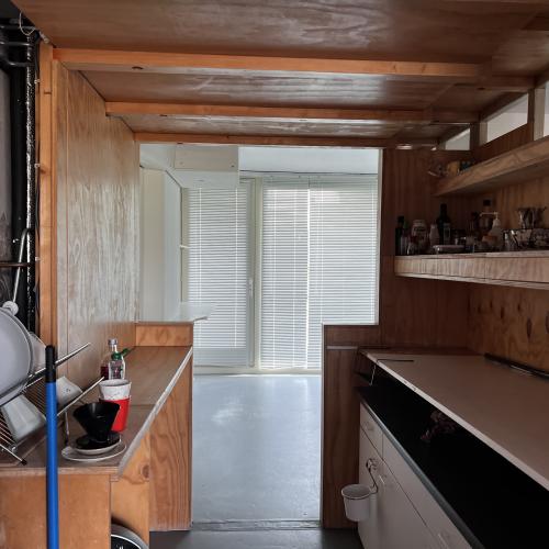 Interieur van een houten bus of caravan: keuken met kastjes, een badkamer en een raam met jaloezieën.