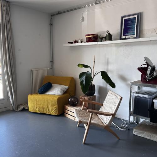 Moderne, lichte woonkamer met een geel bankje, een houten stoel en een wit plankje. Er staat een grote plant en een raam in.