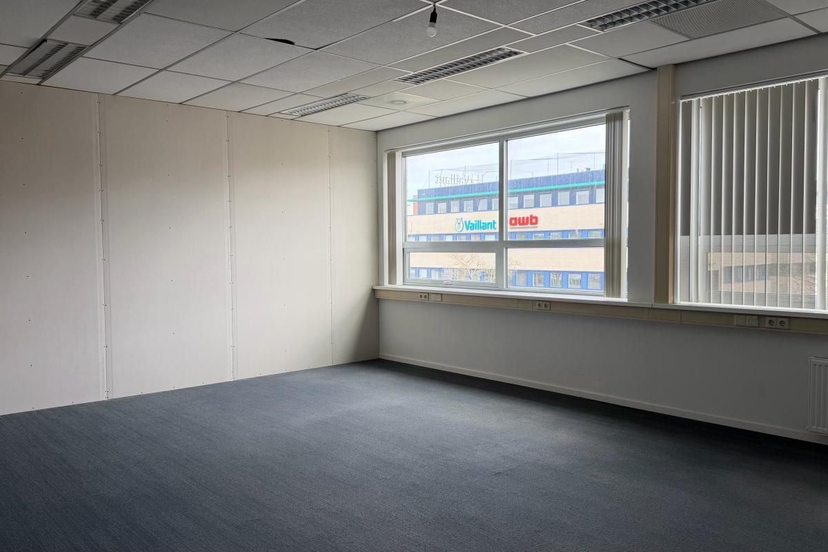 Lege, professionele kamer met grijs vloerkleed, witte muren en grote ramen met jaloezieën.