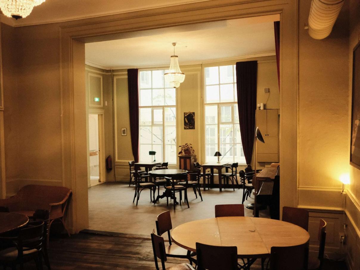 Warm restaurantinterieur met diners en een uitnodigende blik naar een zaleinde met grote ramen.
