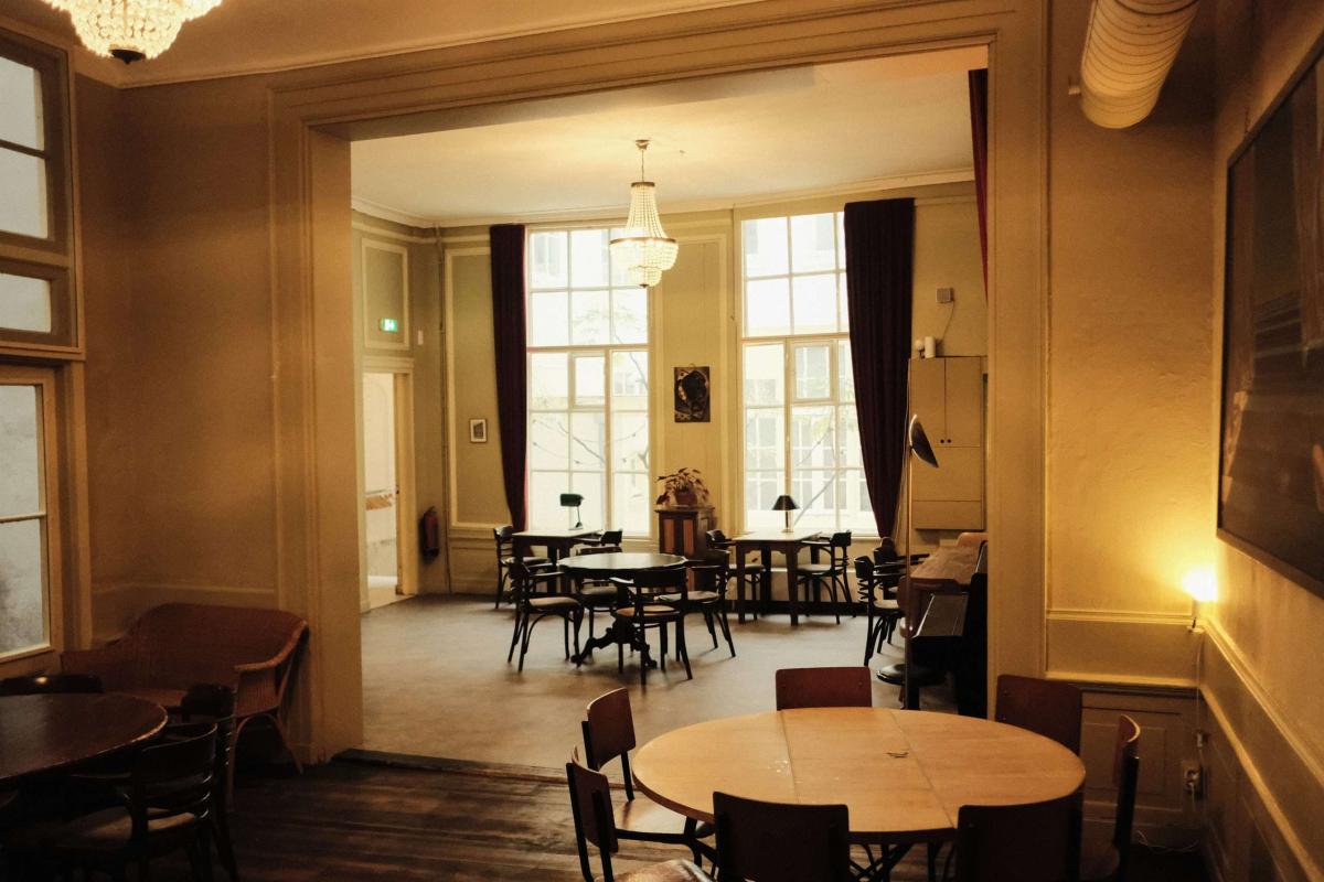 Warm restaurantinterieur met diners en een uitnodigende blik naar een zaleinde met grote ramen.