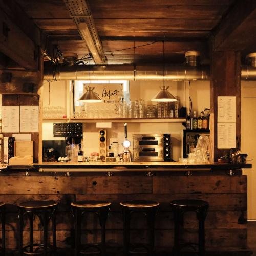 Interieur van een rustiek café: een lange houten bar met planken, verlichting en rijen barstoelen.