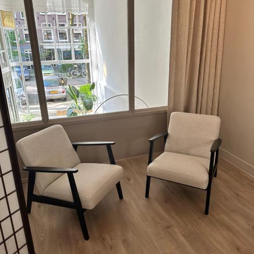 Twee moderne fauteuils met lichte kussens en donker frame staan in een zonnige woonkamer.