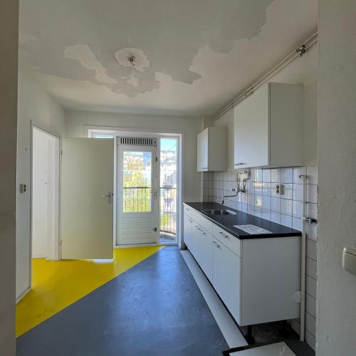 Modern keuken- en badkamergedeelte met witte kasten, grijze vloer en felgeel tapijt op de achtergrond.