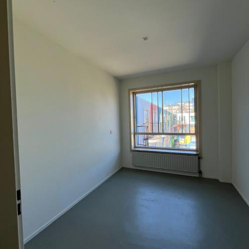 Leeg, modern wooninterieur met witte muren, grijs betonvloer en een raam dat uitzicht geeft op stedelijke gebouwen.