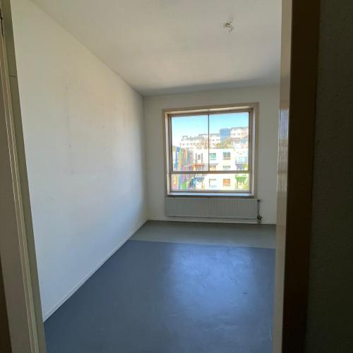 Een lege, moderne kamer met witte muren, een grijs betonvloer en een raam met uitzicht op gebouwen.