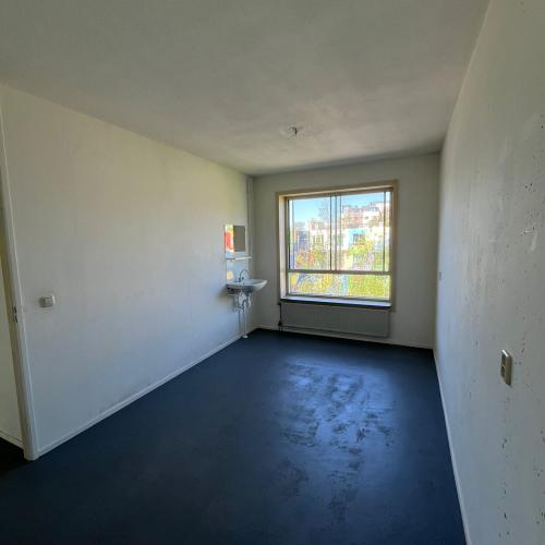 Een lege, lange kamer met lichte muren en donkere vloer, met een raam aan het einde dat daglicht binnenlaat.