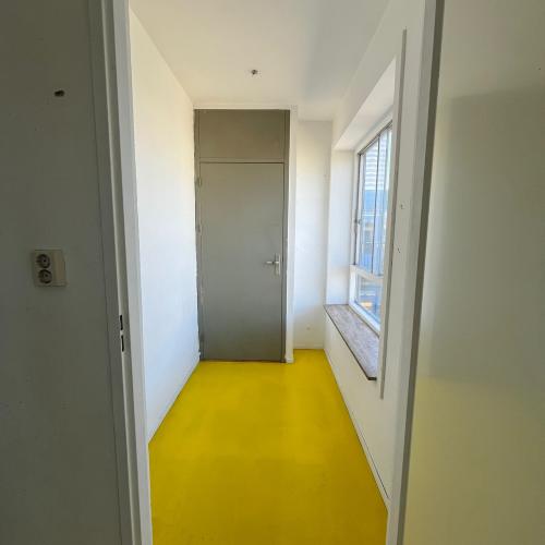Doorgang met helder gele vloer, witte muren en een donkergrijze kast. Rechts is een raam.