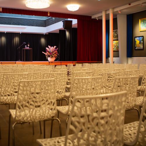Grote zaal met rijen identieke, gele stoelen, gericht naar een podium met rood gordijn.