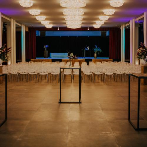 Een grote zaal, opgemaakt voor een evenement, met rijen stoelen, kleine tafels en een podium op de achtergrond.