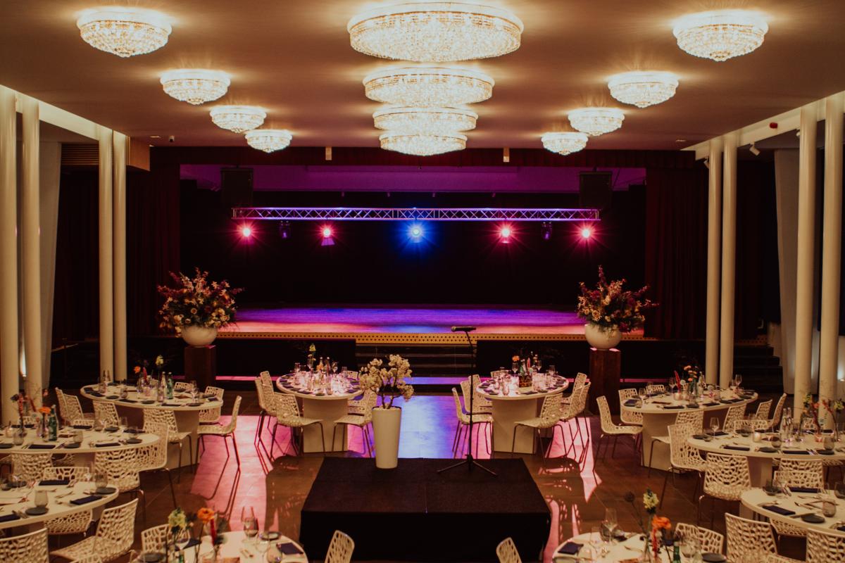 Groot, elegant gehuurd zaal met tafels en stoelen, een podium en grote kroonluchters.