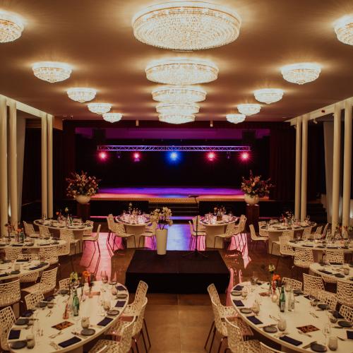 Groot, elegant gehuurd zaal met tafels en stoelen, een podium en grote kroonluchters.