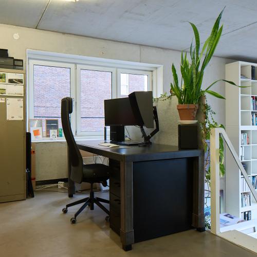 Kantorruimte met modern bureau, twee monitoren, en een boekenplank met boeken en plant.