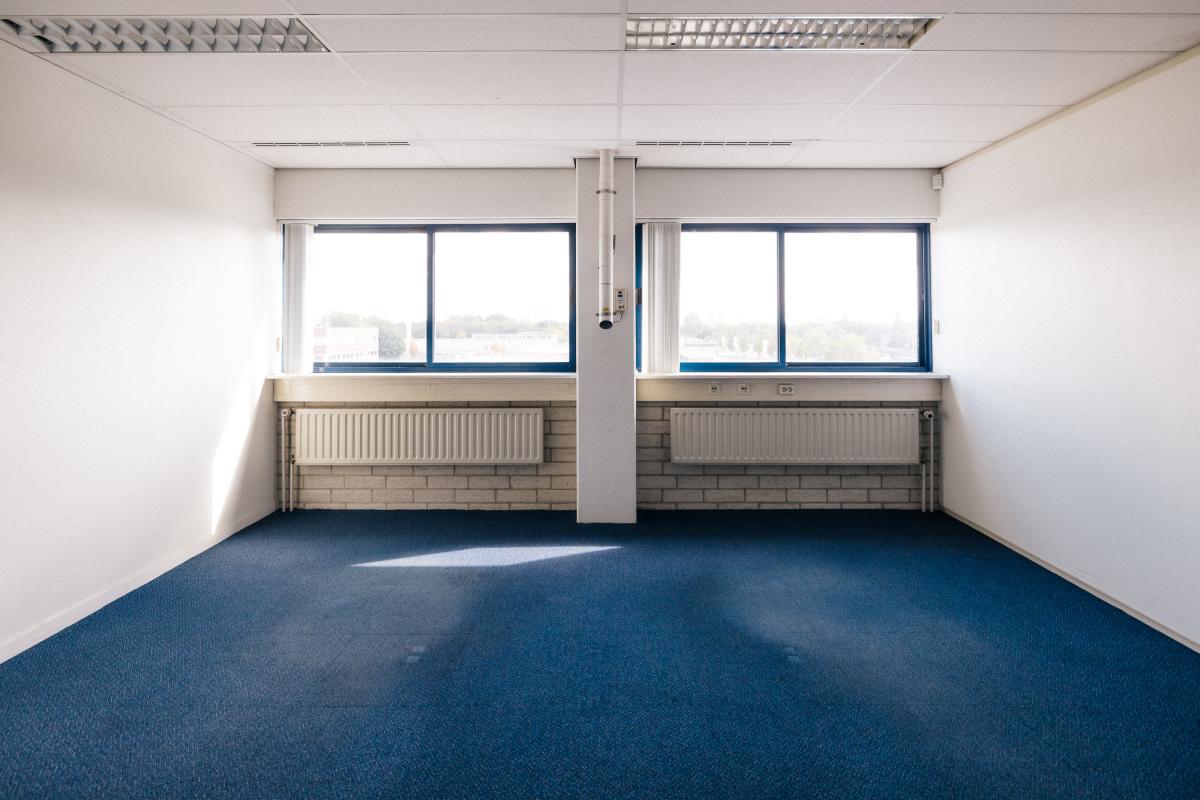 Een leeg, modern kantoor met een blauw tapijt, twee grote ramen en twee radiatoren.