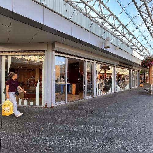 Vrouw loopt met gele tas langs winkelcentrum met glazen gevels en metalen dakconstructie.