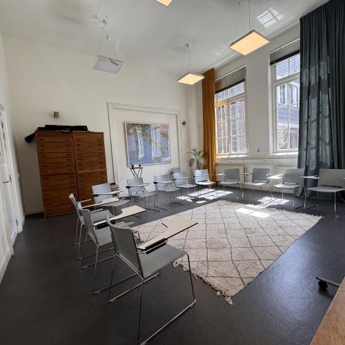 Grote ruimte, stoelen in een kring met klaptafeltjes. Grote archiefkas, ramen met lange gordijnen