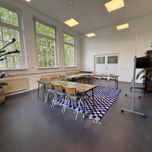 Taalhuis Amsterdam: grote ruimte, tafels met stoelen in u-vorm, whiteboard zij-aanzicht, planten.