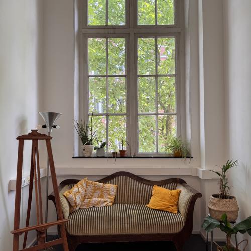 Taalhuis Amsterdam hal: hoog raam met klassiek golvend bankje ervoor. Gele kussens en planten.