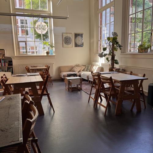 Taalhuis Amsterdam café: 4 tafeltjes met stoelen erom, een hoekbank, hoge ramen, posters aan de muur