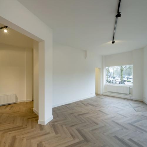 Een lege, lichte kamer met houten parketvloer. Links is een deuropening en rechts grote ramen, wat wijst op een nieuwbouw.