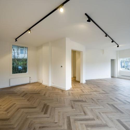 Een lege, moderne kamer met een houten vloer in ruitenvorm. De ruimte is wit en heeft ramen en railsverlichting.